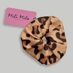 A leopard print satin hair scrunchie with a pink brand tag reading 'Mili Mila Montreal' on a solid gray background.