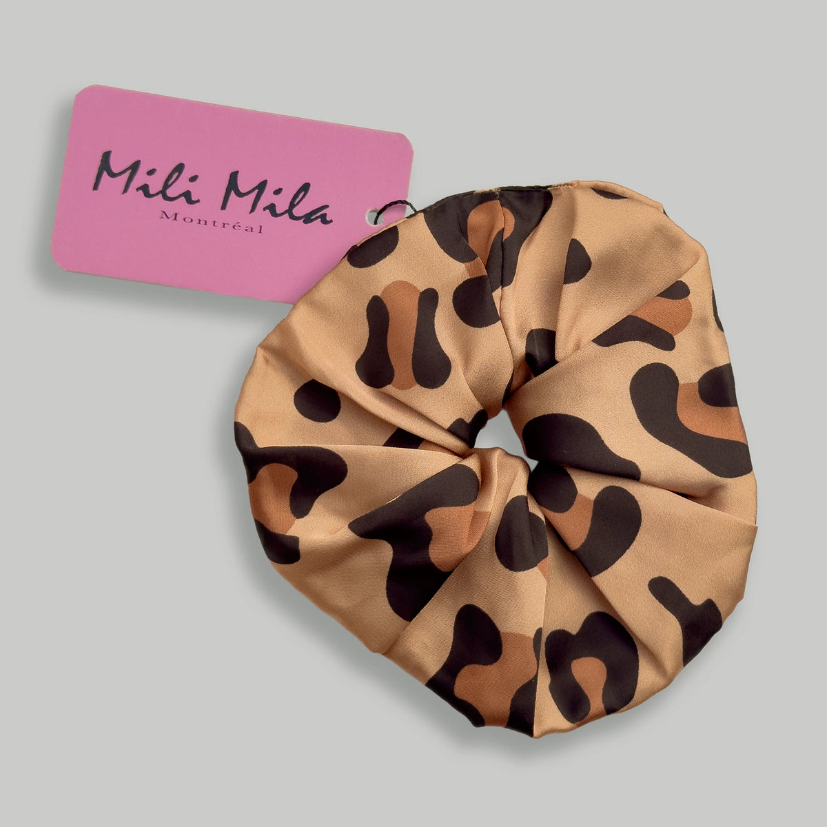 A leopard print satin hair scrunchie with a pink brand tag reading 'Mili Mila Montreal' on a solid gray background.