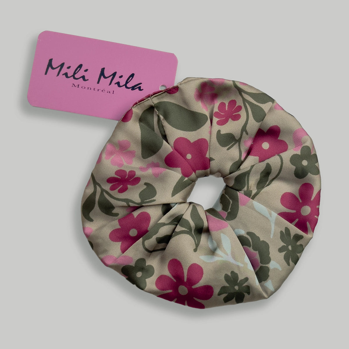 A fabric hair scrunchie with a floral pattern in pink, olive green, and light blue on a beige background, shown with a pink 'Mili Mila Montréal' brand tag on a neutral gray background.